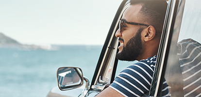 A man with a well-kept beard and sun glasses is leaning out of his parked car to get a good look of the beautiful landscape vista.